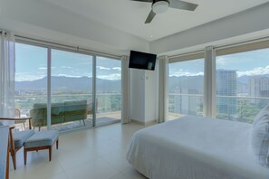 Ein geräumiges Schlafzimmer mit einem großen Bett, einem flachbildfernseher an der Wand und einem Balkon mit Blick auf die Stadt und die Berge.