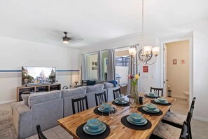 A dining area with a wooden table set for four, a grey sofa, and a television in the background. - Solara Resort - 29170 (Kissimmee)