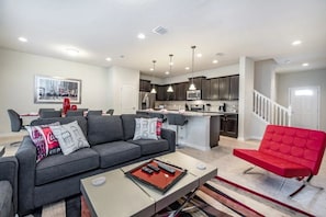 A modern living room with a grey sofa, red chair, and a coffee table with remotes. A kitchen with dark cabinets and a white island is visible in the background. - Windsor at Westside - 2323 (Kissimmee)