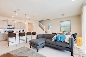 A modern kitchen with a dining area, a gray sectional sofa, and a staircase leading to an upper level. - Windsor at Westside - 3797 (Kissimmee)