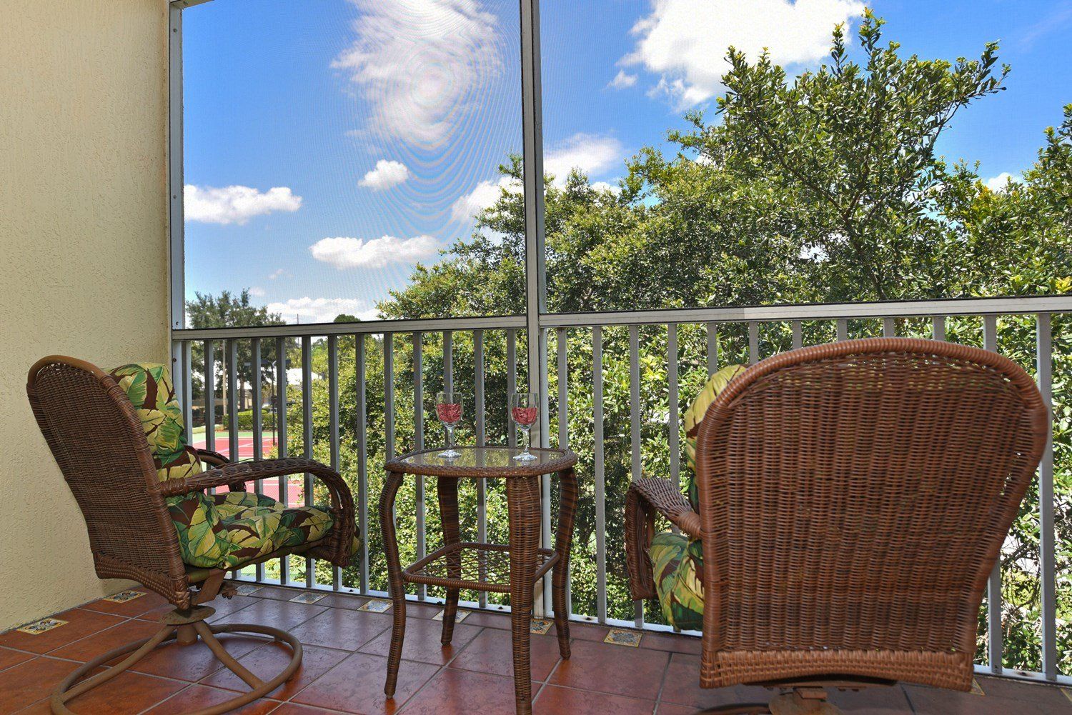 Ein Balkon mit Korbmöbeln, einem Glastisch und Blick auf Bäume und einen klaren Himmel.