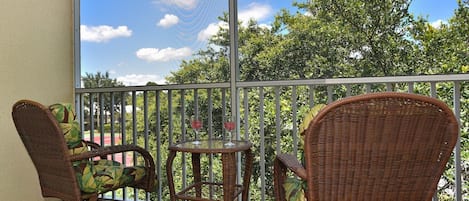 A balcony with wicker furniture, a glass table, and a view of trees and a clear sky.