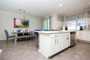 A modern kitchen with a central island, white cabinetry, and a dining area with blue chairs. - Solara Resort - 30486 (Kissimmee)