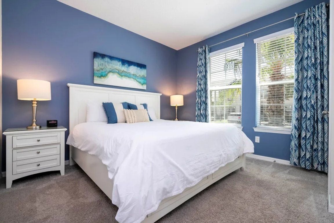A bedroom with a large bed, two bedside tables, a window with blinds, and a blue wall.
