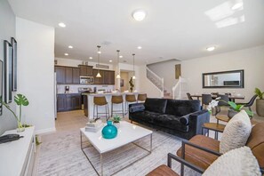 A modern living room with a black sofa, a white coffee table, and a kitchen in the background. - Windsor at Westside - 28836 (Kissimmee)