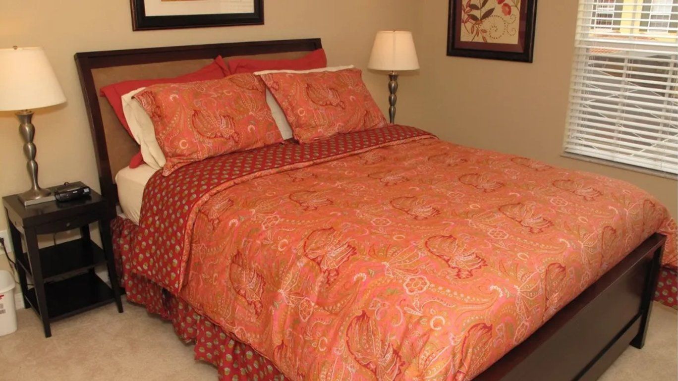 Uma cama com colcha estampada, duas luminárias de cabeceira, um criado-mudo e um quadro na parede.
