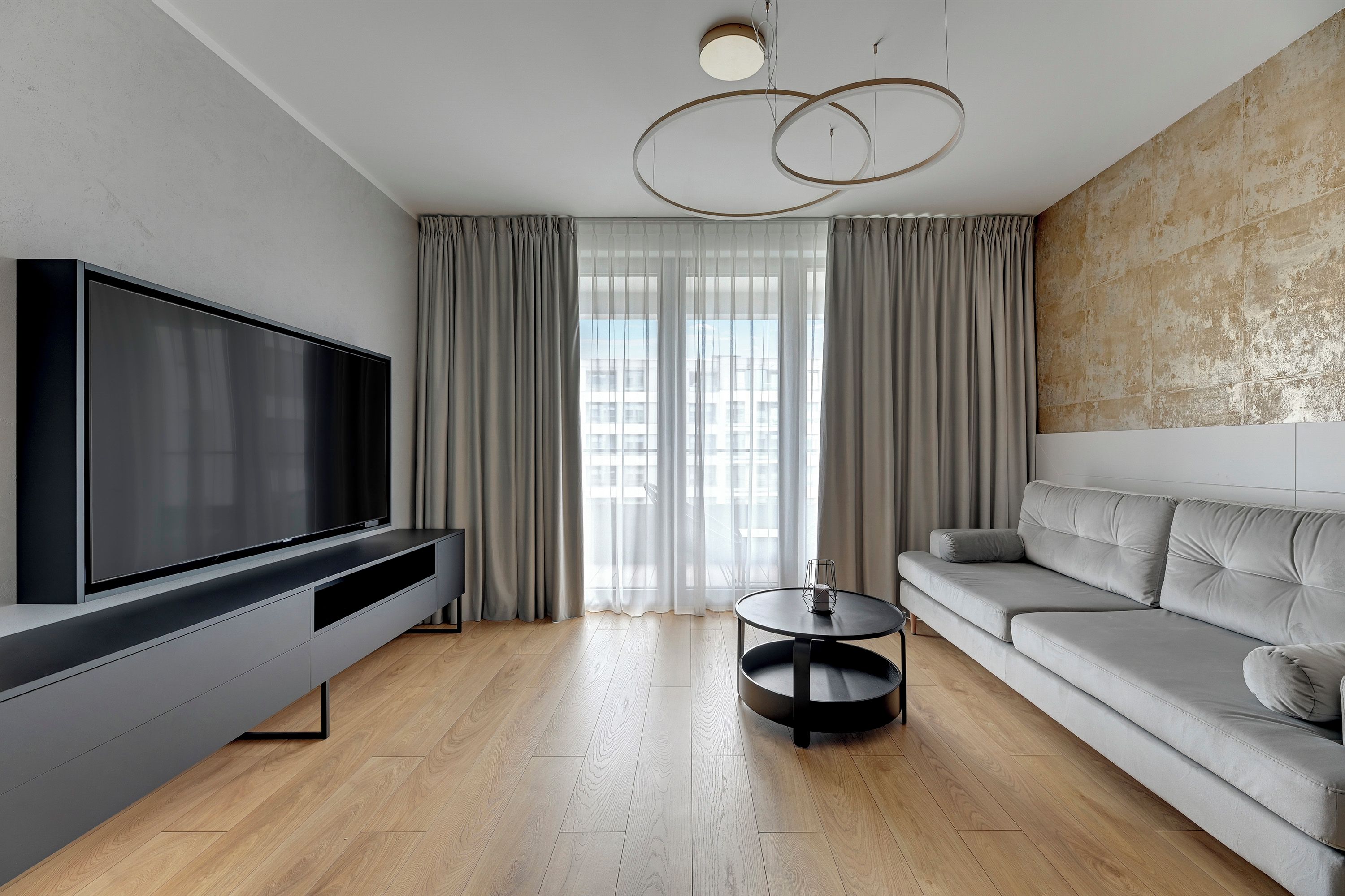 A modern living room with a flat-screen TV, a grey sofa, a round coffee table, and a large window with curtains.