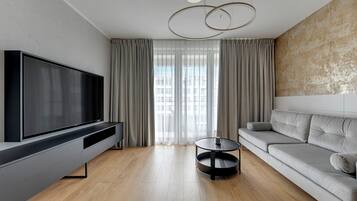 A modern living room with a flat-screen TV, a grey sofa, a round coffee table, and a large window with curtains.