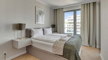 A hotel room with a bed, bedside tables, a lamp, a window with curtains, and a wall-mounted picture.