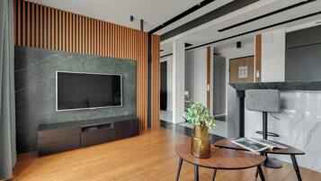 A modern living room with a flat-screen TV mounted on a dark marble wall, wooden paneling, a round coffee table with a vase of green plants, and a magazine.