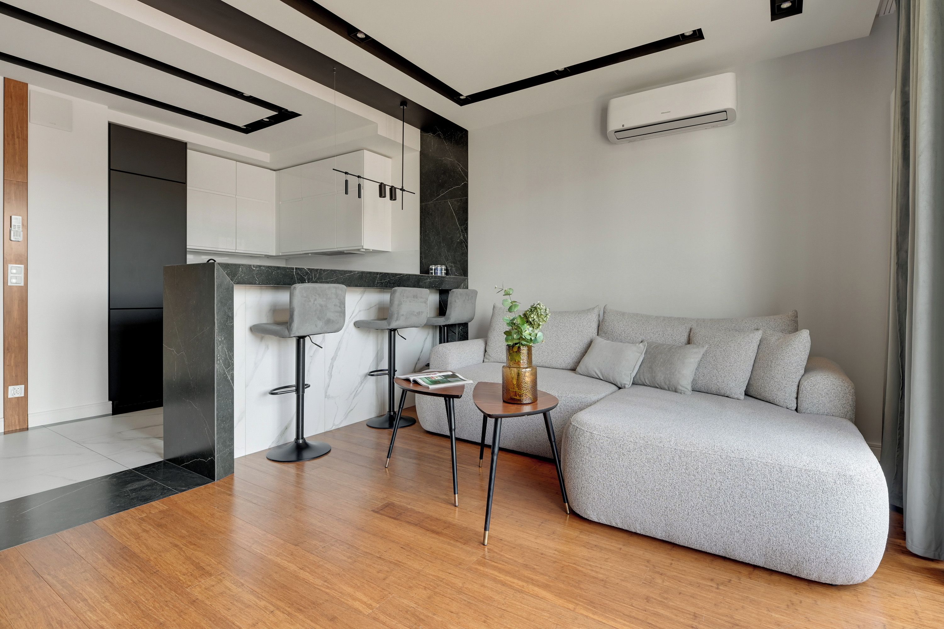 A modern living room with a grey sofa, a small round table with a vase, and a kitchen area with white cabinets and a black countertop.