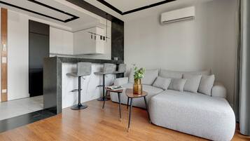 A modern living room with a grey sofa, a small round table with a vase, and a kitchen area with white cabinets and a black countertop.