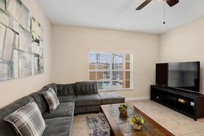 A living room with a sectional sofa, a coffee table with flowers, a flat-screen TV on a stand, and a ceiling fan. - Paradise Palms - 6135 (Kissimmee)