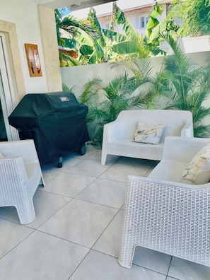 An outdoor patio with a grill, white wicker furniture, and lush tropical plants. - Sun, Pool & Sand! 3BR Family Villa Near the Beach (Cabarete)