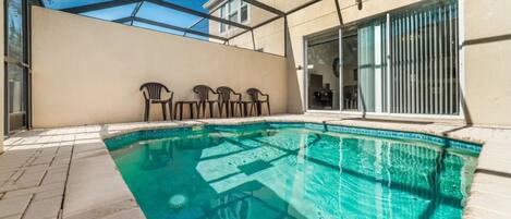 A swimming pool with a glass enclosure, a patio area with chairs, and a sliding glass door leading to an interior space.