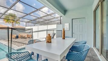 A covered outdoor dining area with a white table and blue chairs, a glass door, and a view of a swimming pool and palm tree.