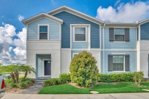 A two-story residential building with a front entrance, a small front yard with a palm tree, and a clear sky. - Windsor at Westside - 7029 (Kissimmee)