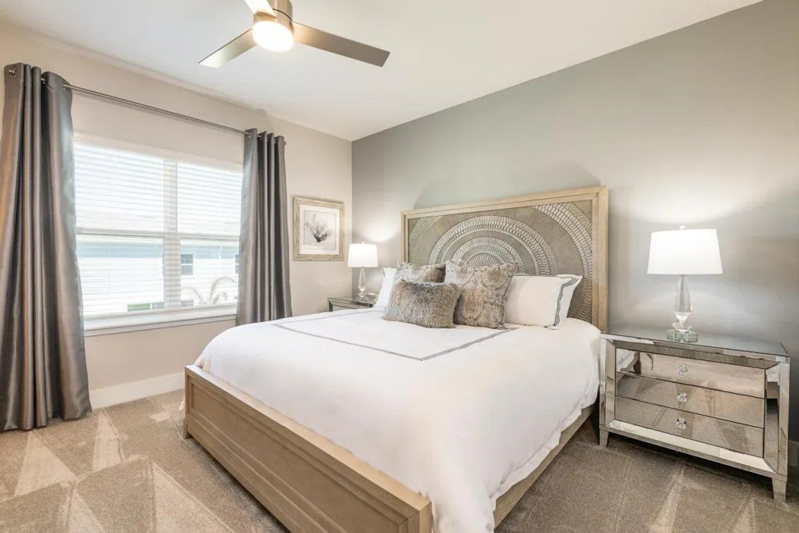 A bedroom with a large bed, a ceiling fan, a nightstand with a lamp, and a window with curtains.