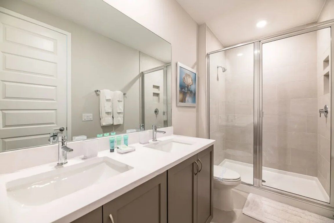 Un baño con tocador doble lavamanos, inodoro y ducha con mampara de vidrio.