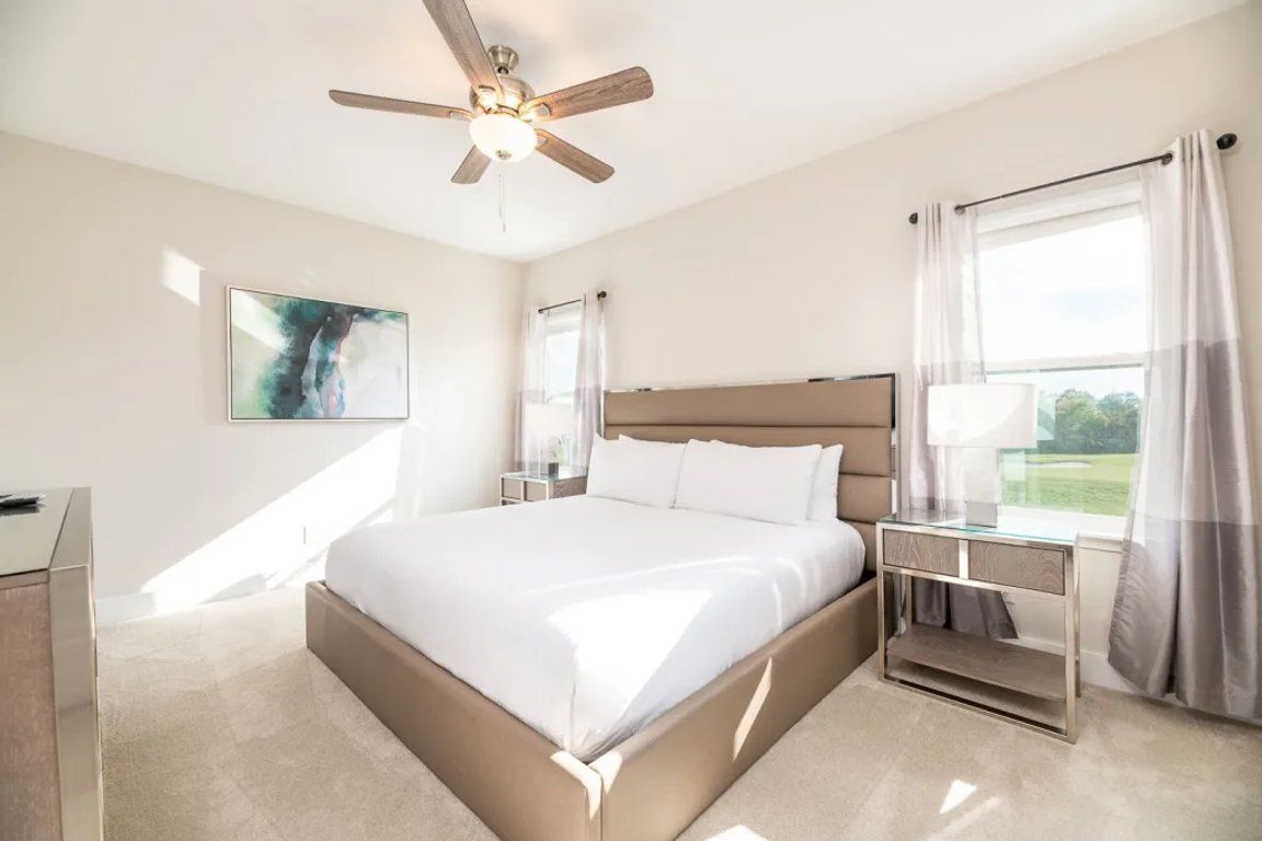 Une chambre d’hôtel moderne avec un grand lit, un ventilateur de plafond, une table de chevet et une fenêtre avec des rideaux.