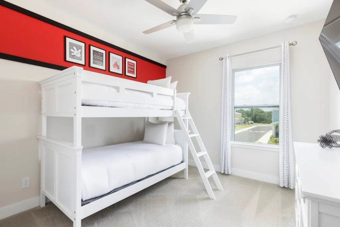 Une chambre avec un lit superposé, un ventilateur de plafond, une fenêtre avec des rideaux et des tableaux encadrés accrochés au mur.