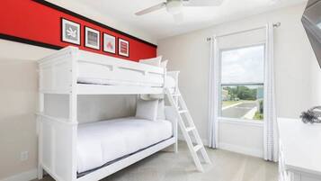 Une chambre avec un lit superposé, un ventilateur de plafond, une fenêtre avec des rideaux et des tableaux encadrés accrochés au mur.