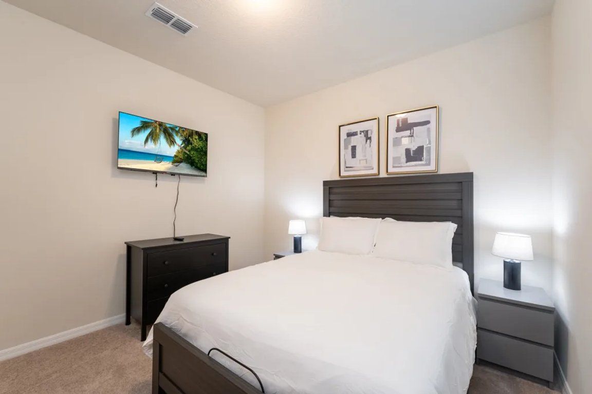 Un dormitorio con cama, mesita de noche con lámpara, cómoda y televisor montado en la pared.