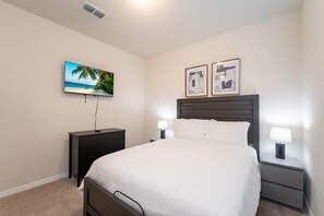 Un dormitorio con cama, mesita de noche con lámpara, cómoda y televisor montado en la pared.