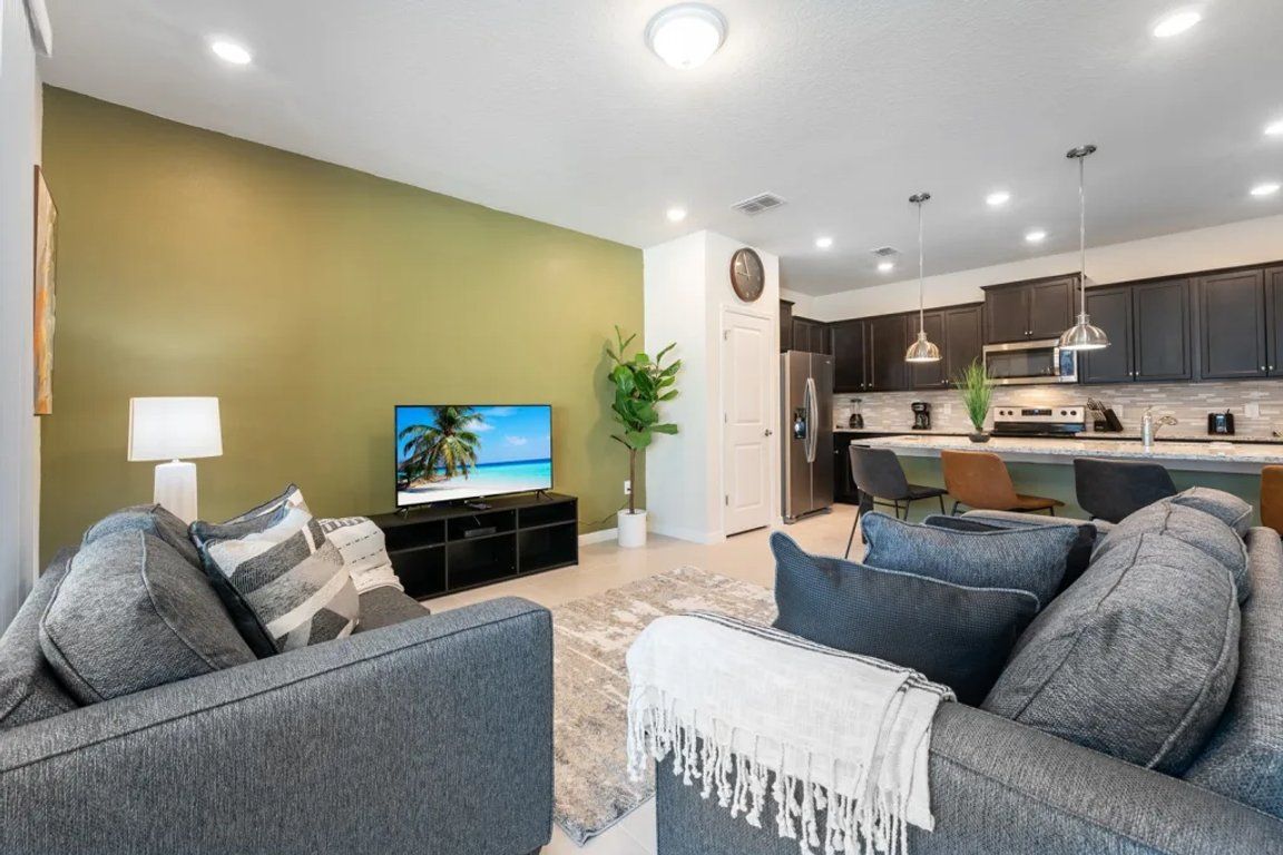 A modern living room with a flat-screen TV, grey sofas, a coffee table, and a kitchen in the background.