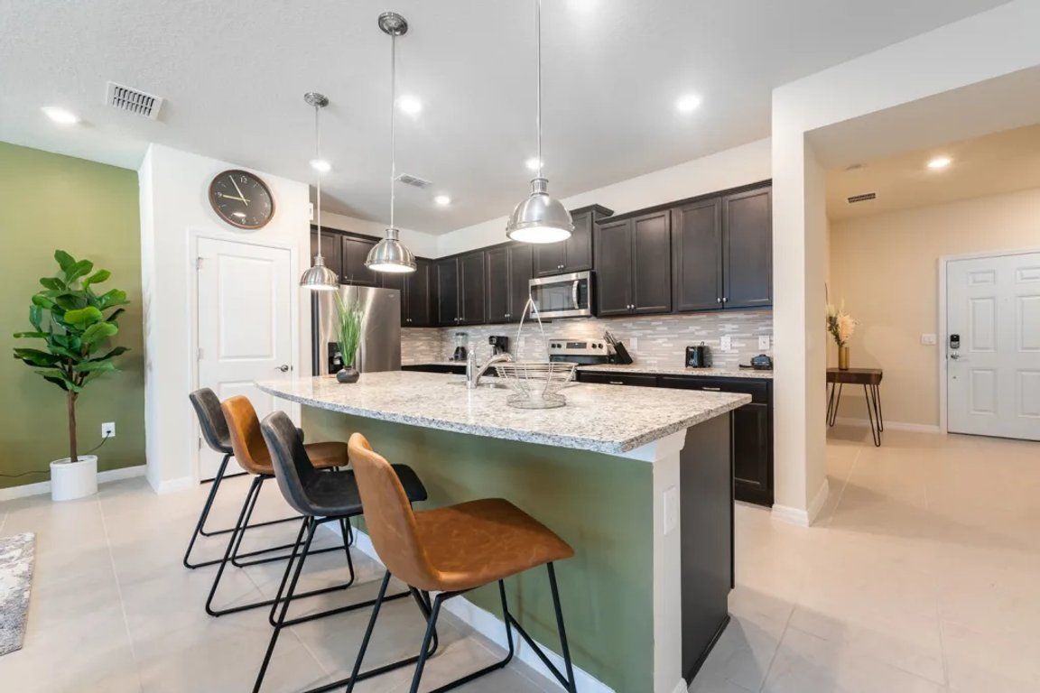 A modern kitchen with dark cabinets, a central island with bar stools, pendant lighting, and a clock on the wall.