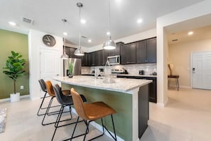 A modern kitchen with dark cabinets, a central island with bar stools, pendant lighting, and a clock on the wall. - Windsor Island - 7711 (Davenport)