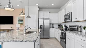A modern kitchen with white cabinetry, stainless steel appliances, granite countertops, pendant lighting, and a flat-screen TV mounted on the wall.