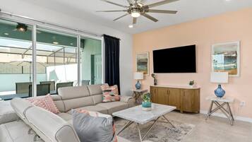 A modern living room with a ceiling fan, a flat-screen TV, a sofa set, a coffee table, and a view through a glass door.