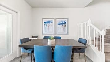 Un comedor con una mesa rectangular, seis sillas azules, dos cuadros abstractos enmarcados en la pared y una escalera con barandillas blancas.