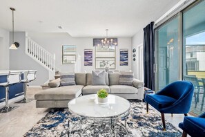 A modern living room with a sofa, a coffee table, and a blue chair. There is a staircase with white railings and a chandelier. - Windsor at Westside - 7899 (Kissimmee)