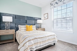 A bedroom with a bed, two bedside tables, a window with blinds, and a wall-mounted artwork. - Windsor at Westside - 7899 (Kissimmee)