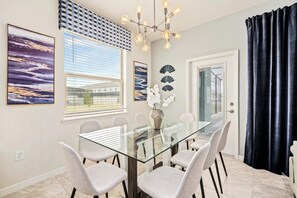 A dining area with a glass table, six chairs, a chandelier, and artwork on the walls. - Windsor at Westside - 7899 (Kissimmee)