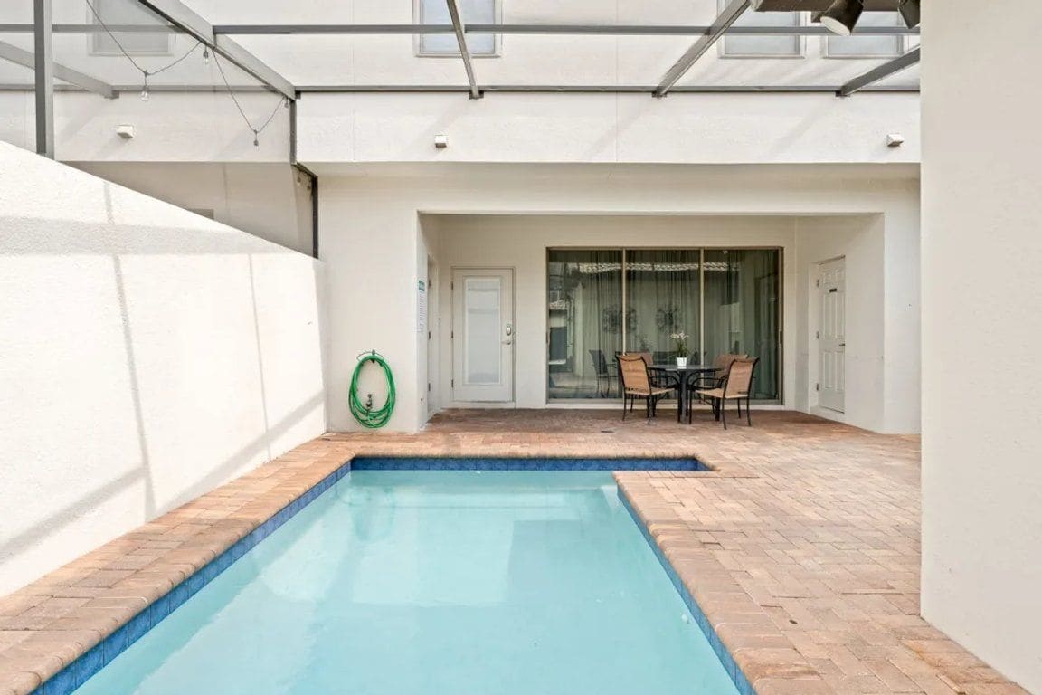 An outdoor swimming pool with a tiled deck, a table and chairs, and a glass door leading to a covered area.