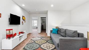 A modern living room with a grey sofa, a flat-screen TV mounted on the wall, and a white entertainment unit with toys.