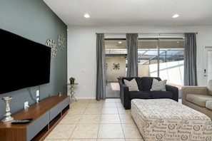 A modern living room with a flat-screen TV, a sofa, a coffee table, and a patterned ottoman. - Windsor at Westside - 8106 (Kissimmee)