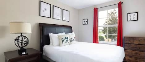 A bedroom with a bed, a nightstand, a dresser, a ceiling fan, and a window with red curtains.
