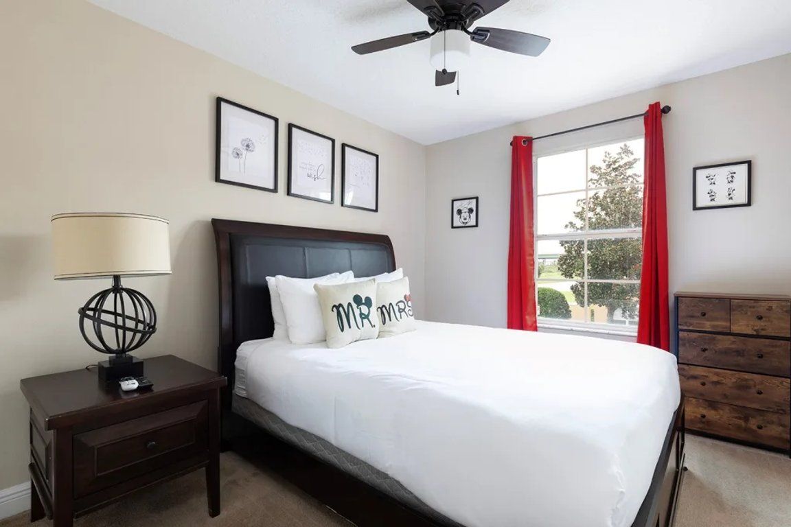 A bedroom with a bed, a nightstand, a dresser, a ceiling fan, and a window with red curtains.
