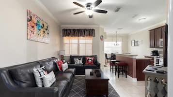 A living room with a black leather sofa, a coffee table, a ceiling fan, and a kitchen area with a microwave and a stove.