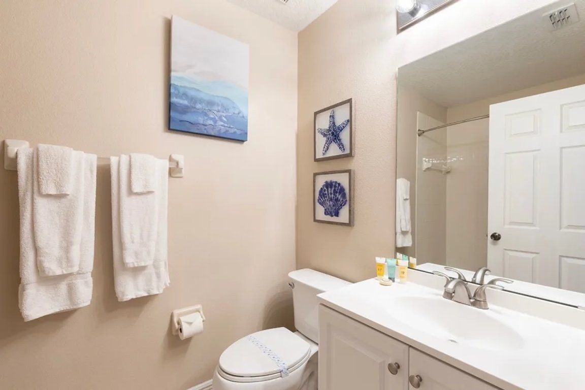 Une salle de bain avec des toilettes, un lavabo et un miroir. Des serviettes sont accrochées au mur et un tableau encadré est placé au-dessus des toilettes.