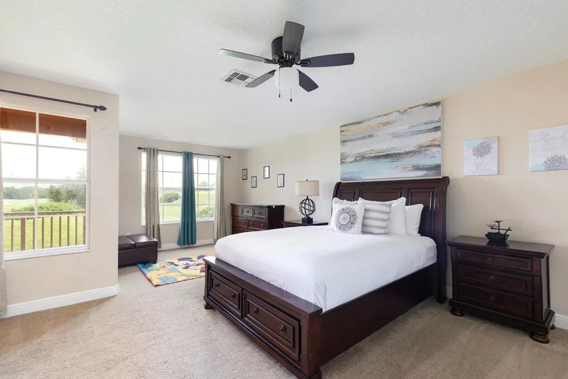 A bedroom with a large bed, a ceiling fan, a window with curtains, and a view of the outdoors.