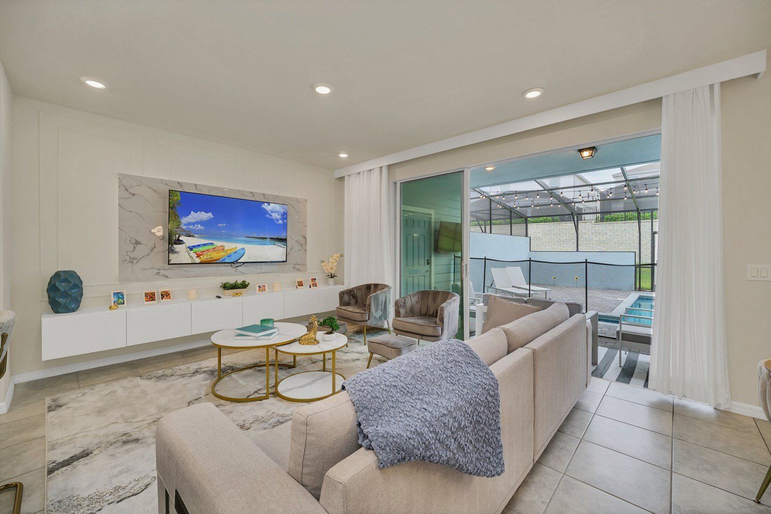 A modern living room with a large TV, comfortable seating, a glass door leading to a balcony with a pool.