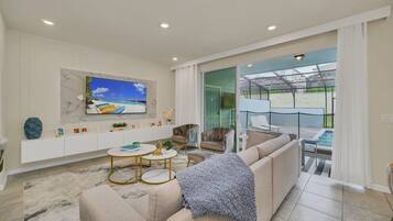 A modern living room with a large TV, comfortable seating, a glass door leading to a balcony with a pool.