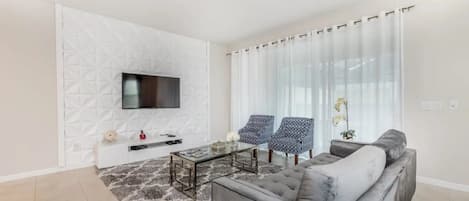 A modern living room with a sofa, two armchairs, a coffee table, a flat-screen TV mounted on a textured wall, and a large window with sheer curtains.