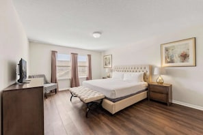 A hotel room with a large bed, a bedside table, a lamp, a bench, a TV, and a window with curtains. - Windsor at Westside - 25863 (Kissimmee)