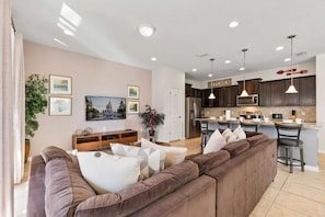 A modern living room with a large brown sectional sofa, a kitchen area with dark cabinets and a bar, and a dining area with a small table and chairs. - Windsor at Westside - 25863 (Kissimmee)